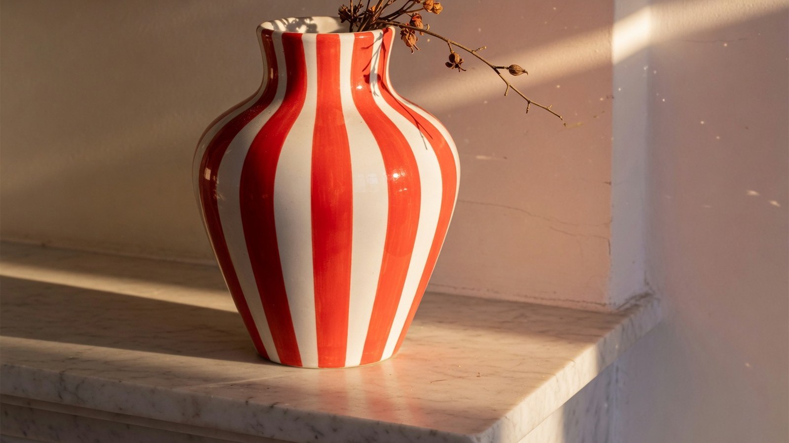 Vase en céramique blanc et rouge rayé, posé sur une étagère en marbre, éclairé par la lumière latérale.