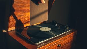 Meuble hi-fi noyer années 1960, couvercle ouvert, main posant disque vinyle sur tourne-disque, lumière dorée.