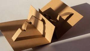 Découpe en carton kraft se dépliant en plis géométriques nets, texture ondulée visible et ombres graphiques, éclairage dramat