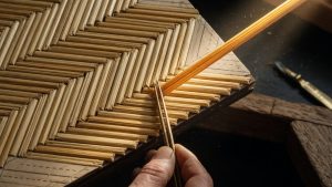 Main à la pince posant un brin de paille dorée dans un panneau de marqueterie au motif chevron