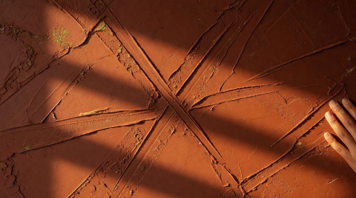 hero-1 Gros plan sur une surface d'enduit à la chaux teint terracotta, éclairage rasant révélant la texture et les reliefs du minera
