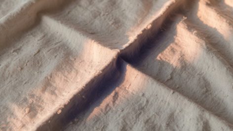 Gros plan d'une main froissant un panneau de lin lavé en lumière rasante, révélant les irrégularités et plis du tissu en tons