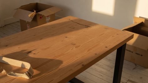 Bureau en bois brut avec grain visible, fissures et rayures, entouré de cartons de déménagement ouverts dans la lumière de fi